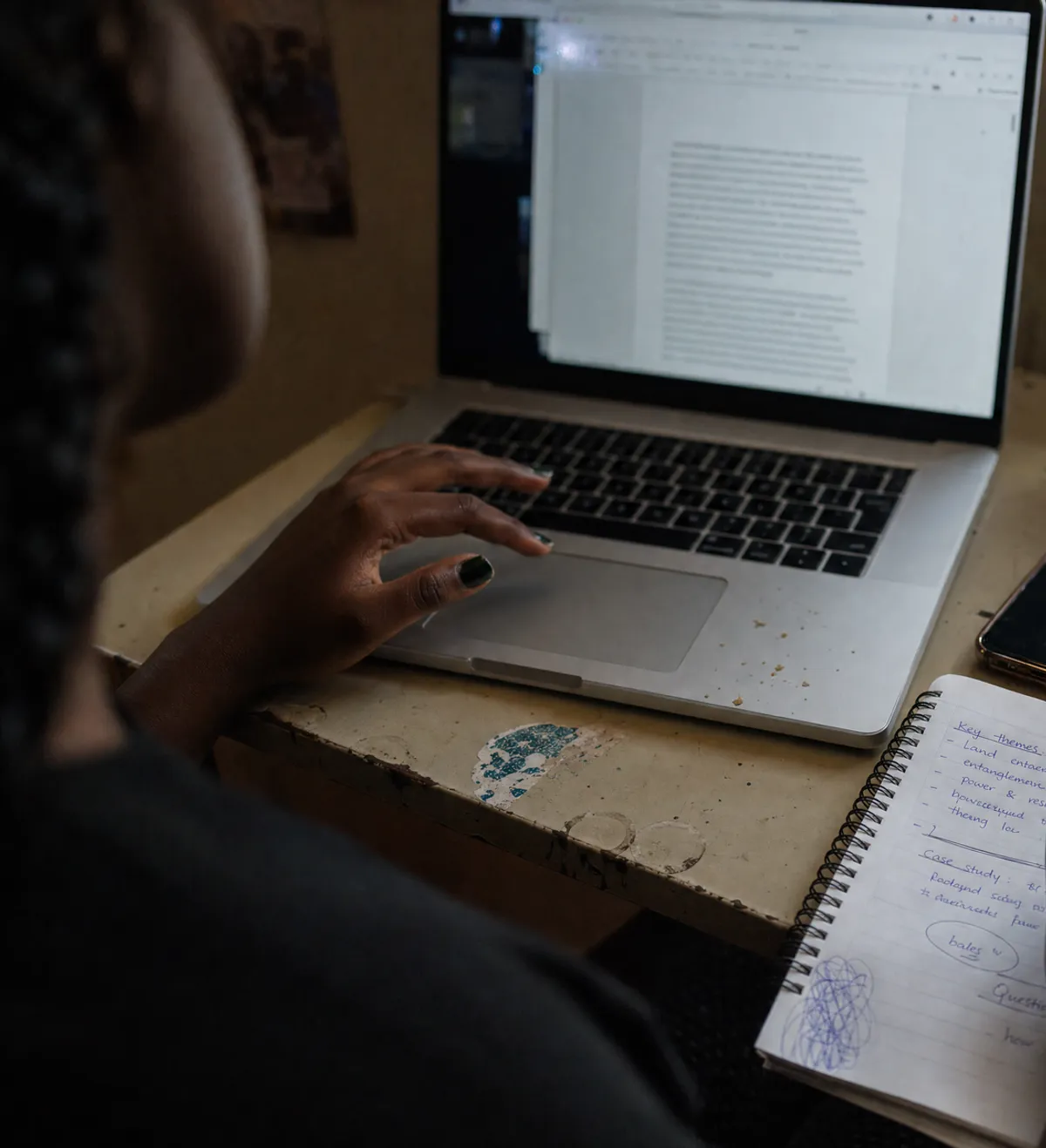 A hand writing notes in a spiral notebook beside a laptop showing a document