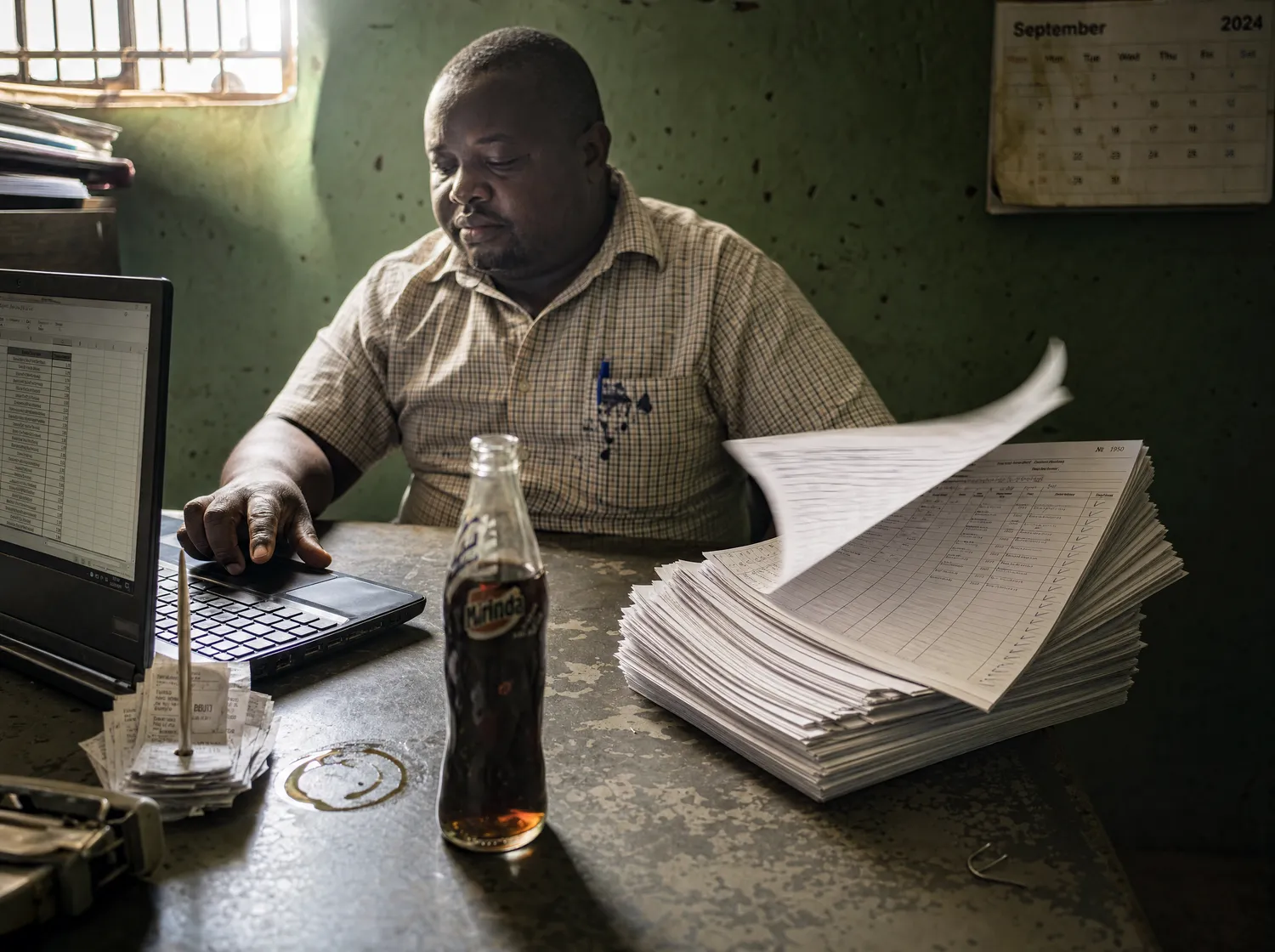 A person at a laptop next to a tall stack of paper forms in a small office