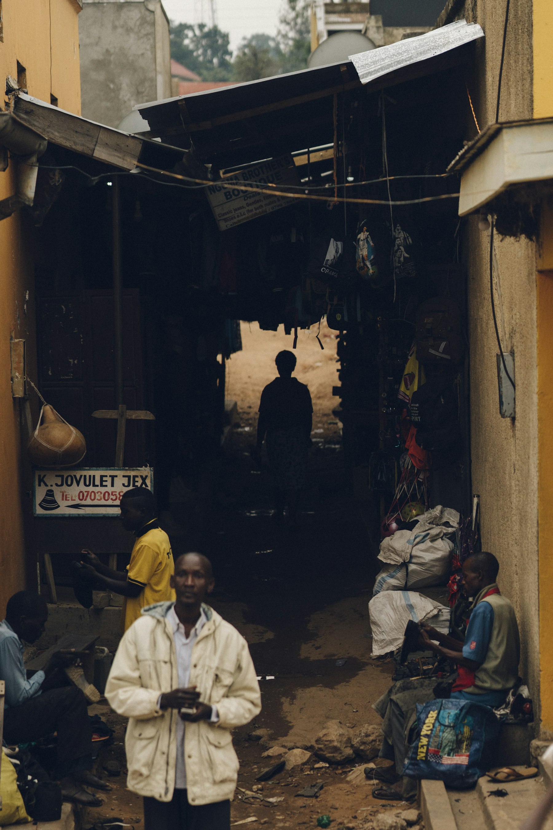 A narrow market alley in Uganda, with traders and a cobbler at work